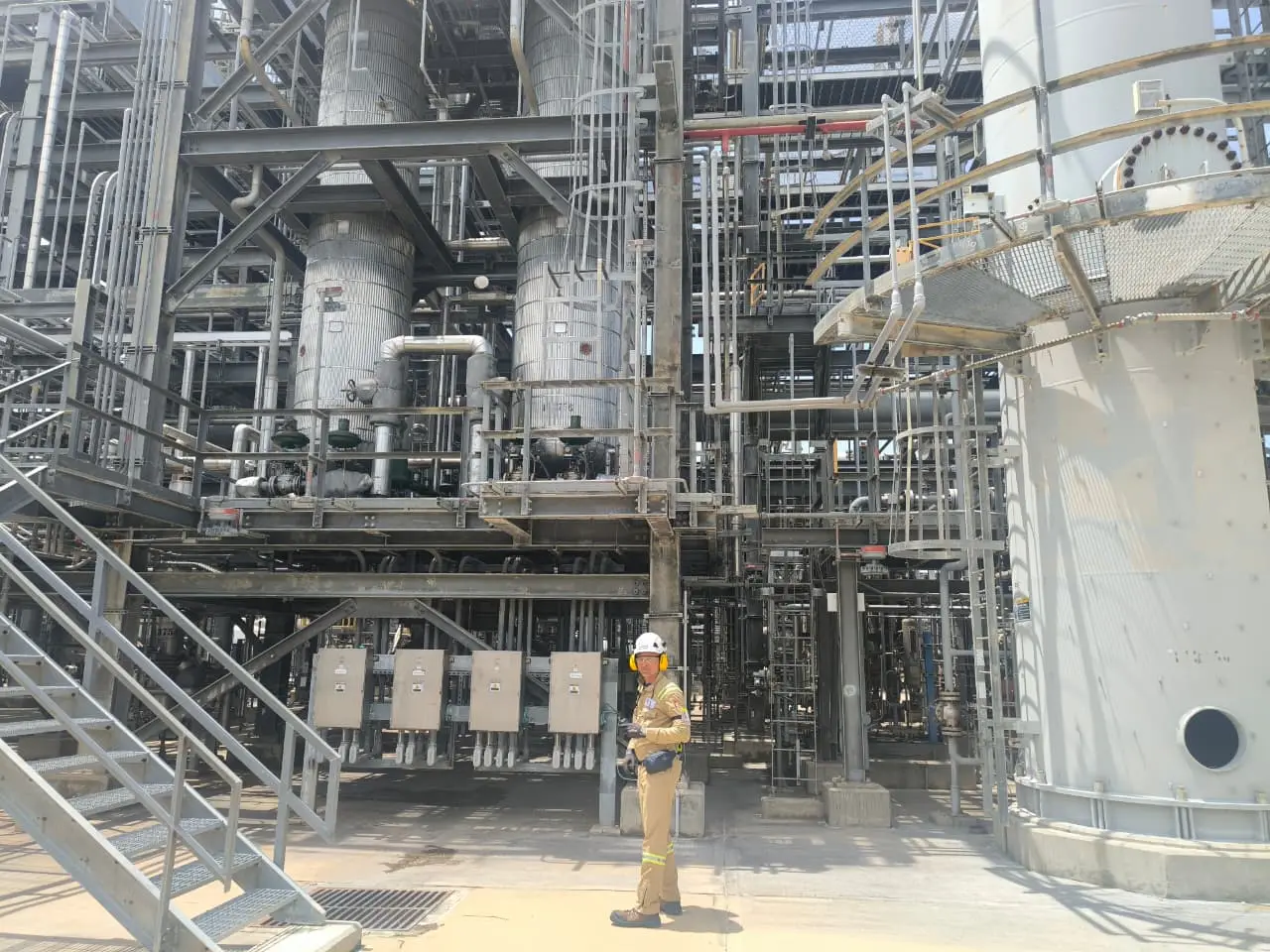 Profesional con casco junto a equipos de proceso, tuberías y estructuras metálicas industriales