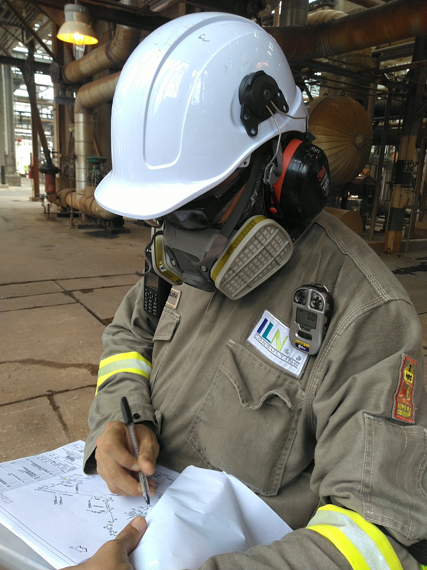 Profesional de ILN con casco y protección respiratoria registra información en un plano durante una revisión en campo